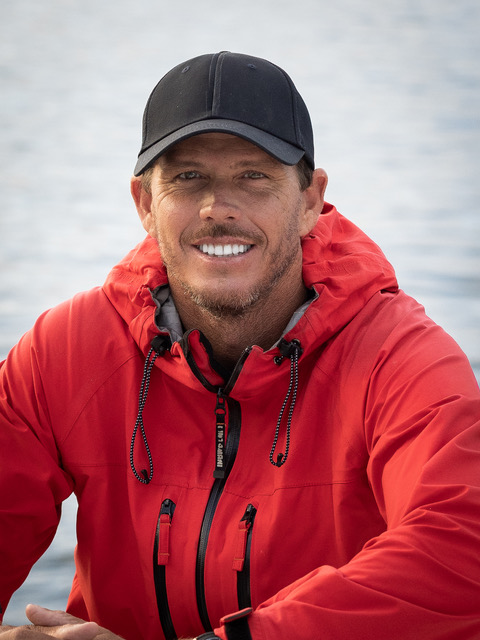 Mike Coots smiling, wearing a black cap and red jacket, with calm water behind him.